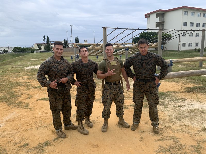 Emmanuel Colmenares in a plate carrier flashing a shaka, flanked by three Marines in camouflage utilities at an obstacle course in Okinawa.