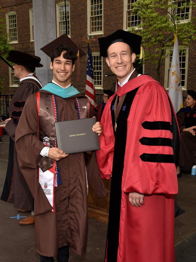 Emmanuel Colmenares in a brown Brown University gown, MPA hood, and Watson School stole, holding his diploma cover beside Dean John N. Friedman in red doctoral regalia, University Hall behind them.