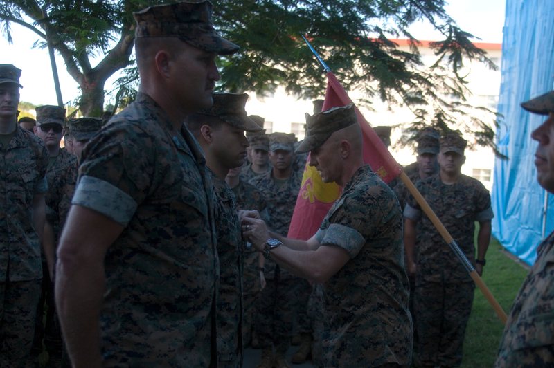 Sergeant Emmanuel Colmenares standing at attention in camouflage utilities as Colonel Robert B. Brodie pins a medal to his chest, with Marines in formation and the 31st MEU colors behind them.