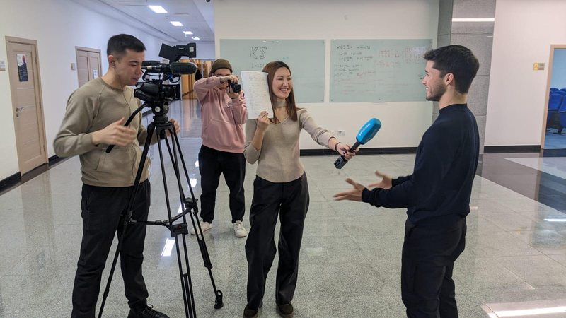 Emmanuel Colmenares being interviewed by a Qazaqstan TV reporter holding a blue microphone, with a camera operator and photographer, in a hallway at Nazarbayev University.