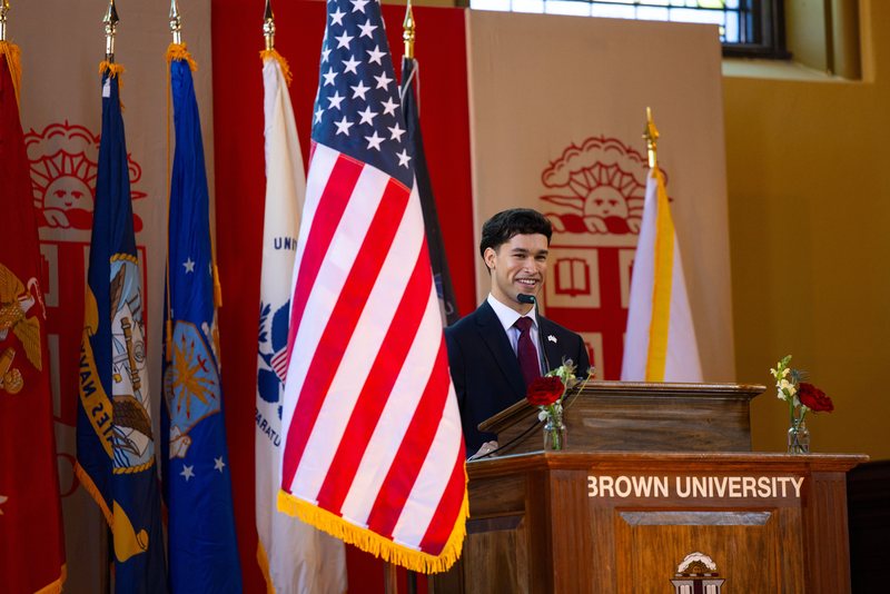Emmanuel Colmenares smiling at a Brown University lectern in Sayles Hall, in a dark suit and burgundy tie, with the United States and service flags behind him.
