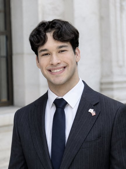 Emmanuel Colmenares, in a pinstriped suit and navy tie, smiling in front of a marble colonnade.